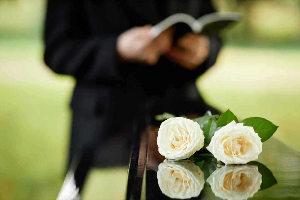 Nahaufnahme eines Sargs, darauf liegen zwei weiße Rosen. Im Hintergrund ein Trauerredner