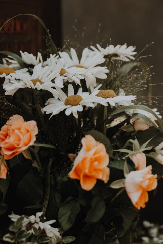 Ein Strauß mit weißen Gänseblümchen mit gelber Mitte, umgeben von orangefarbenen Rosen und grünem Blattwerk, sanft arrangiert in sanftem, natürlichem Licht. Perfekt, um während einer Beerdigung in Dresden Trost zu spenden.