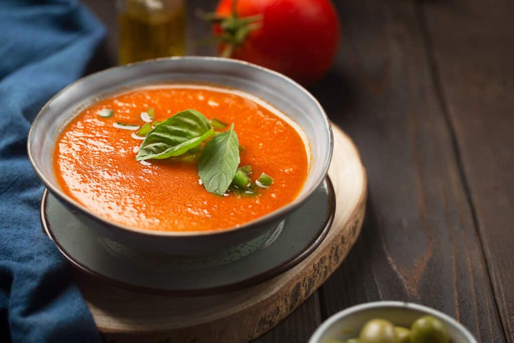 Eine Schüssel Tomatensuppe, garniert mit frischen Basilikumblättern und gehackten Frühlingszwiebeln, steht auf einem Holztisch und erinnert an die gemütlichen Mahlzeiten, die bei Familientreffen eingenommen wurden. Im Hintergrund sind ein blaues Tuch, eine Tomate und eine kleine Flasche Olivenöl zu sehen, die die Wärme der Bestattung Dresden-Veranstaltungen widerspiegeln.