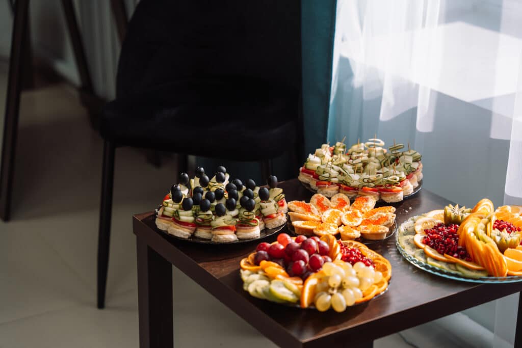 Auf einem Holztisch wird eine Auswahl an Snacks präsentiert, darunter Obstteller mit Trauben, Orangen und Granatäpfeln sowie verschiedene Vorspeisen, garniert mit schwarzen Oliven, Gurkenscheiben und rotem Kaviar. Sonnenlicht fällt durch ein nahes Fenster in den Empfangsbereich der Bestattung Dresden.