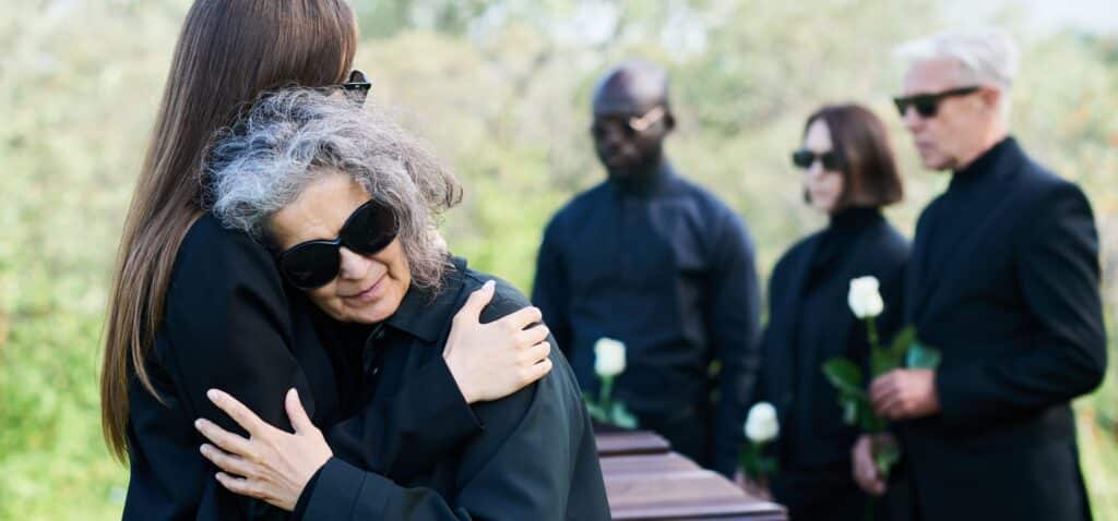 Mehrere Personen in schwarzer Kleidung stehen trauernd um einen Sarg. Im Vordergrund umarmen sich zwei Frauen emotional, während andere Gäste mit weißen Rosen im Hintergrund Abschied nehmen. Die Szene findet im Freien statt und symbolisiert den Moment des Verlusts eines geliebten Menschen.
Das Bild verdeutlicht die emotionale Tragweite von Vorsorge – insbesondere die Wichtigkeit, frühzeitig ein Testament zu schreiben. Ein rechtssicheres Testament-Muster kann helfen, Angehörige in solchen Momenten vor zusätzlicher Belastung zu schützen und den Nachlass klar zu regeln.