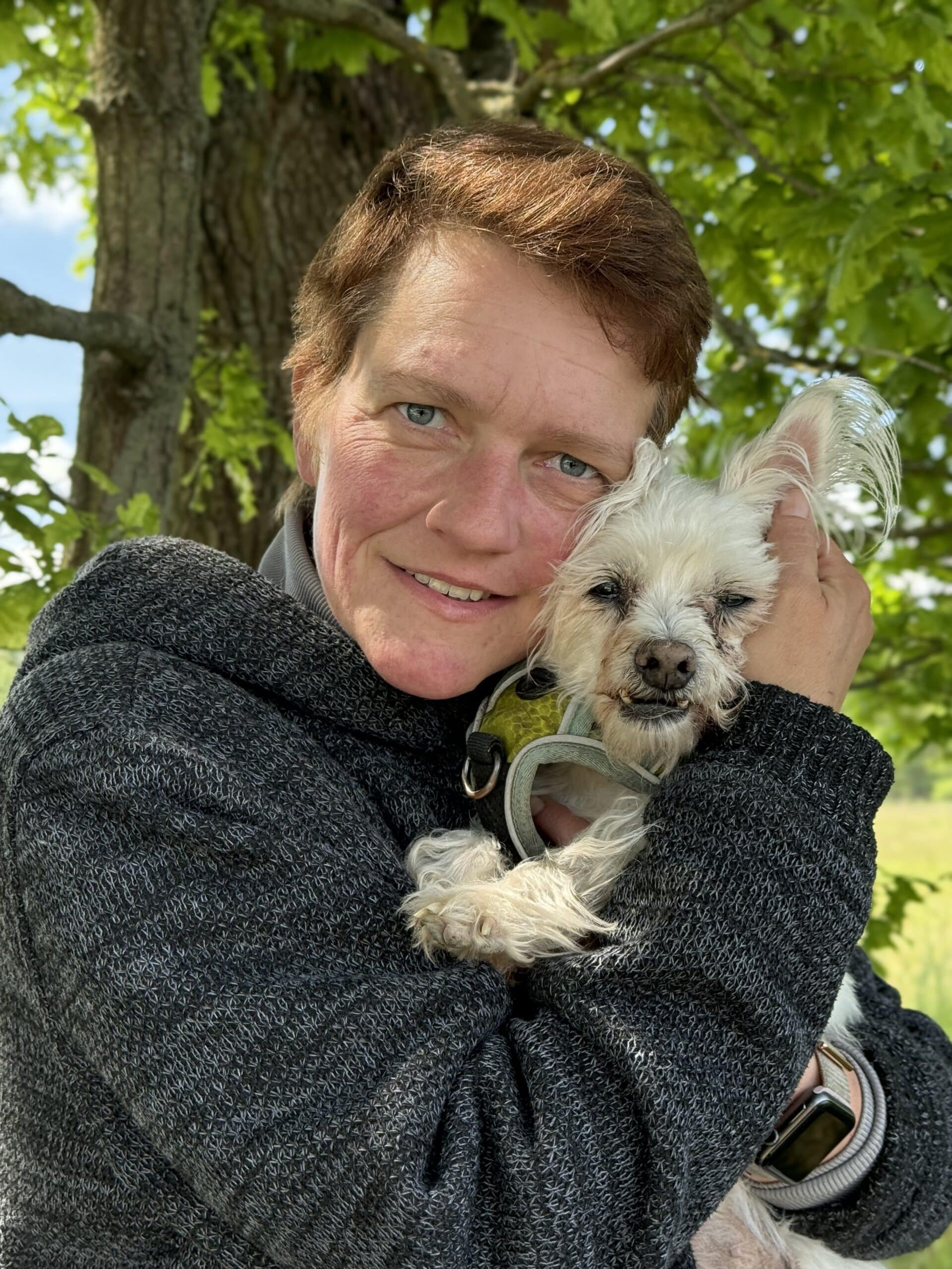 Eine Person mit kurzen Haaren, die einen grauen Pullover trägt, lächelt warm, während sie im Freien vor einem Baum mit grünen Blättern steht und einen kleinen weißen Hund umarmt.