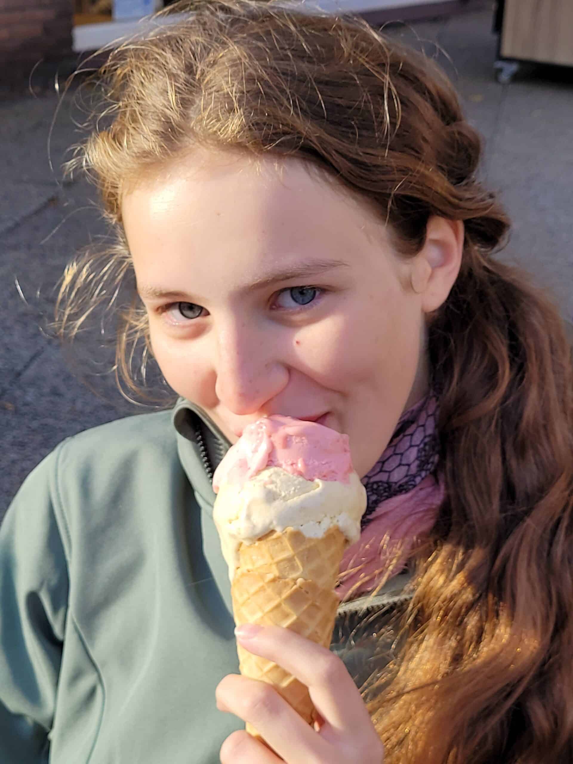 Eine junge Person mit langen braunen Haaren genießt eine große Eistüte mit rosa und weißen Kugeln, lächelt und schaut in die Kamera bei natürlichem Außenlicht.