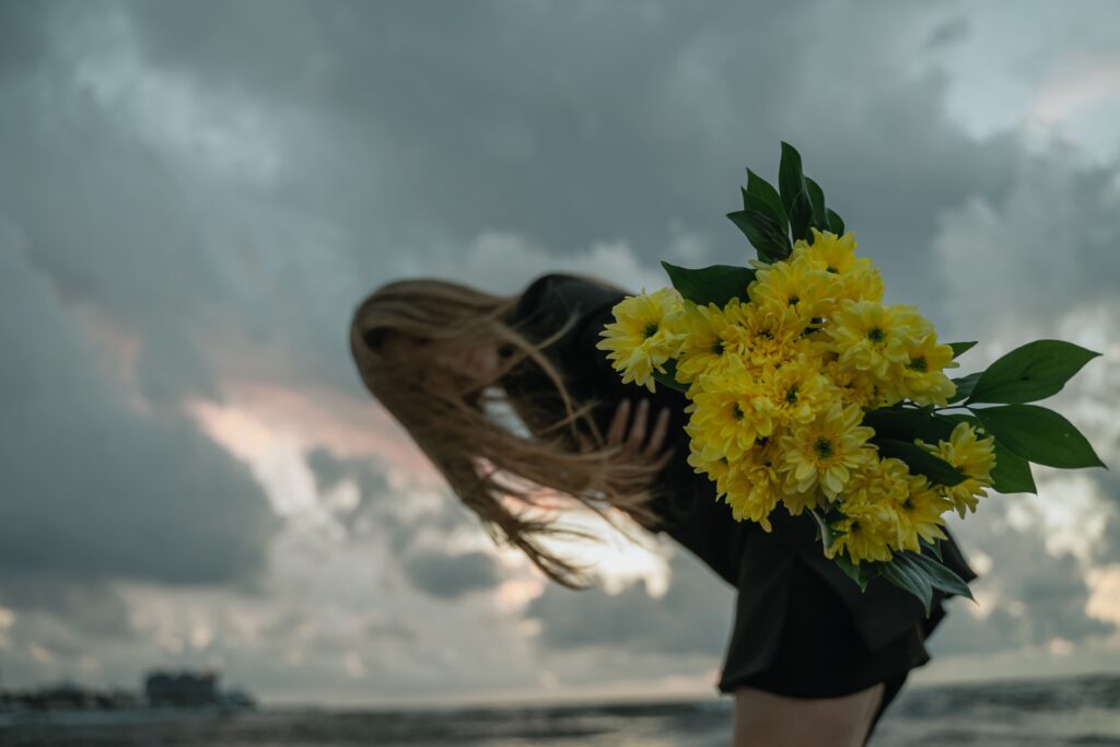 Eine schwarz gekleidete Person mit langen Haaren steht unter einem wolkenverhangenen Himmel am Wasser. Der Wind weht ihr die Haare ins Gesicht, während sie einen großen Strauß gelber Blumen in der Hand hält – ein stiller Moment des Abschieds, wie gemacht für Trauersprüche zur Seefahrt.