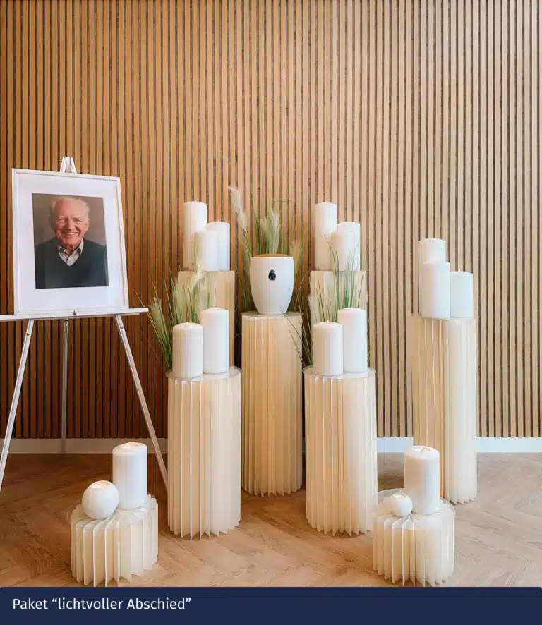 Eine Gedenkstätte mit großen weißen Kerzen, einer weißen Urne, grünen grasähnlichen Pflanzen und dem gerahmten Foto eines älteren Mannes, die auf cremefarbenen Sockeln vor einer Wand mit Holzlatten angeordnet sind, hebt günstige Bestattungen ab 1600 Euro hervor.