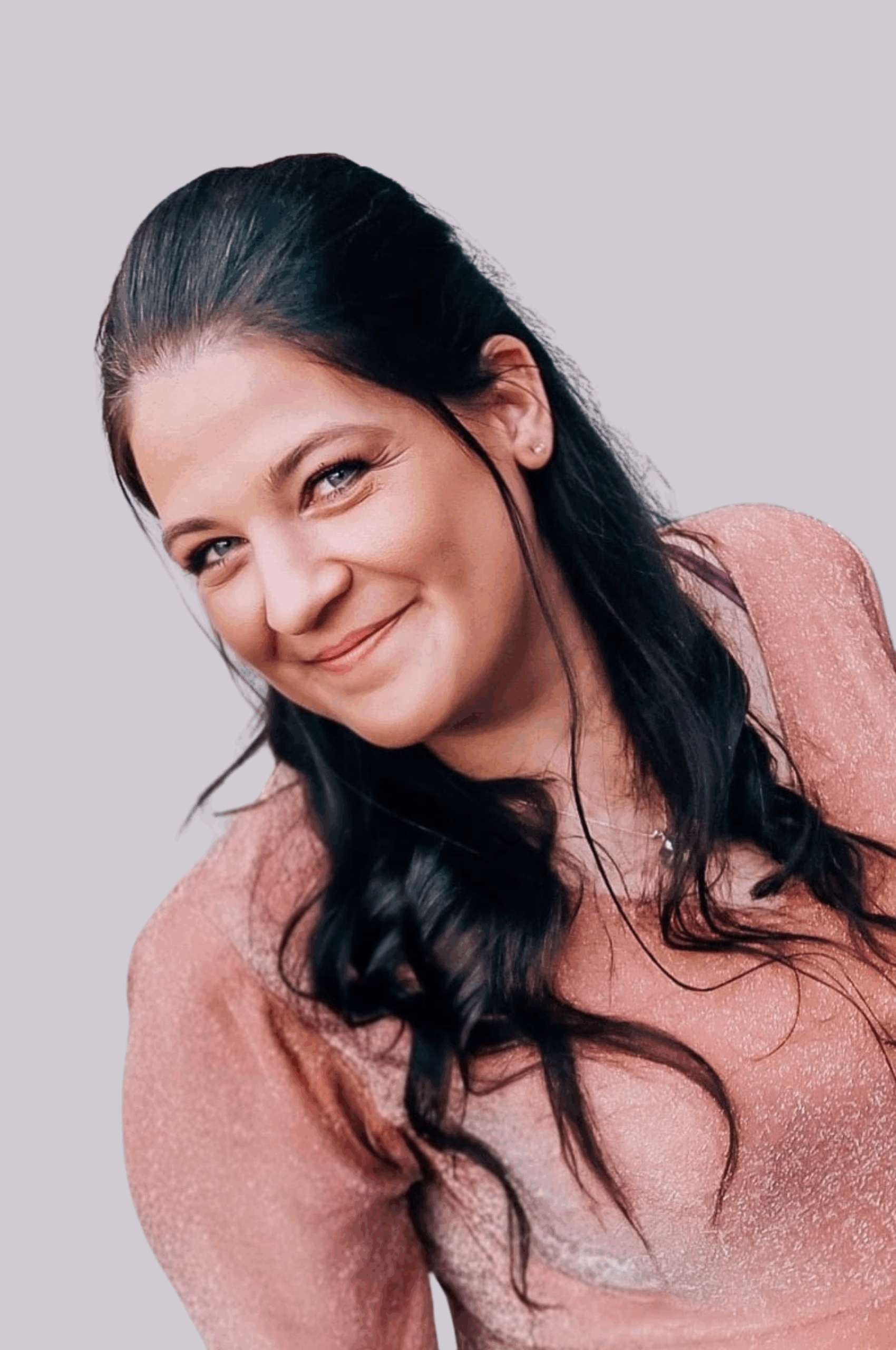 Eine Frau mit langem, dunklem, gewelltem Haar und heller Haut lächelt sanft. Sie trägt ein schimmerndes rosa Oberteil und steht vor einem schlichten, hellen Hintergrund.