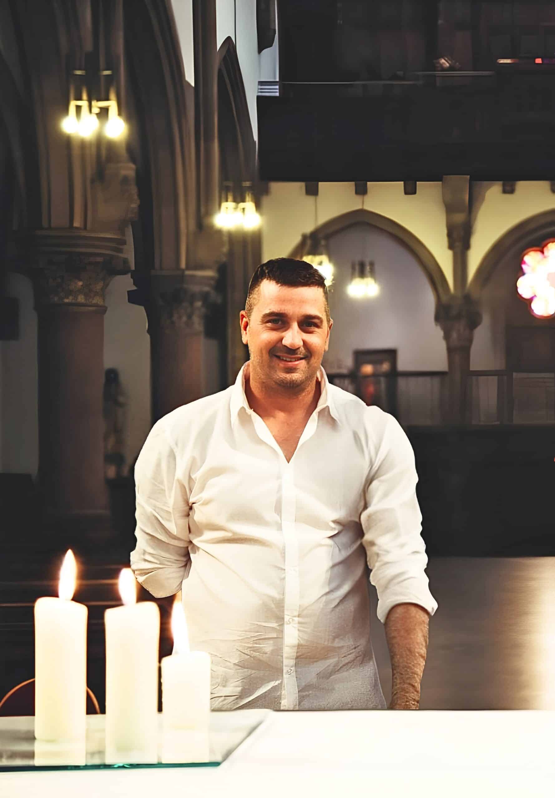Ein Mann in einem weißen Hemd steht lächelnd neben einem Tisch mit drei brennenden Kerzen in einer schummrigen Kirche, hinter ihm sind Gewölbedecken und verzierte Säulen zu sehen.